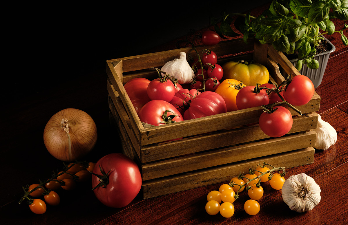 An assortment of various tomatoes in a wooden crate with garlic and fresh basil.