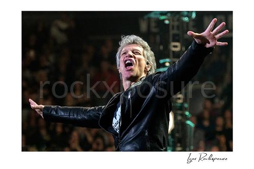 Horizontal color image of Jon Bon Jovi in a dramatic live performance close-up, singing with both arms outstretched and the arena crowd blurred behind him