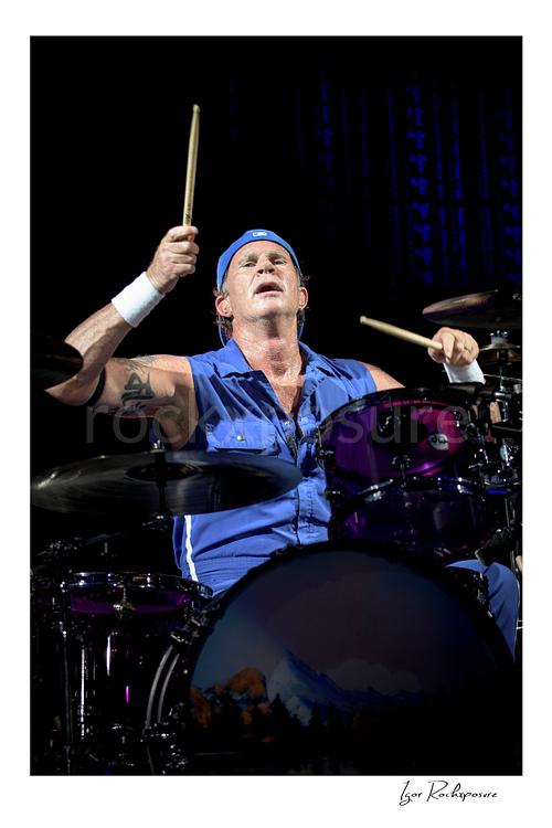 Vertical color image of Chad Smith of Red Hot Chili Peppers at the drum kit in a dramatic live performance portrait, raising both drumsticks above a purple drum set against a dark stage backdrop