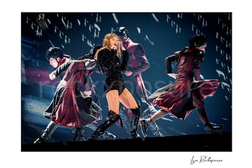 Horizontal color image of Taylor Swift singing on stage with dancers in sweeping coats under dramatic concert lighting