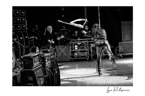 Horizontal black and white image of Yngwie Malmsteen tossing his guitar across the stage toward legendary guitar tech Thomas Nordegg during a live performance, captured in a dynamic wide stage moment