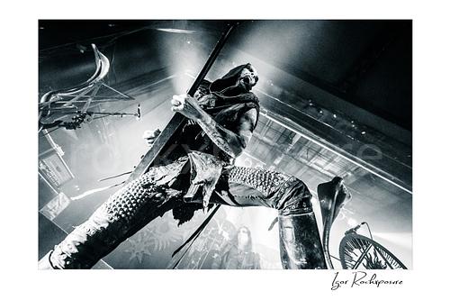 Horizontal black and white image of Nergal of Behemoth performing with a guitar in a dramatic low-angle stage shot under intense backlight