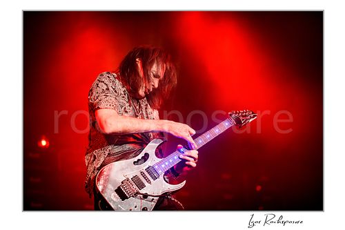 Horizontal color image of Steve Vai playing guitar under vivid red stage lighting with glowing fret markers
