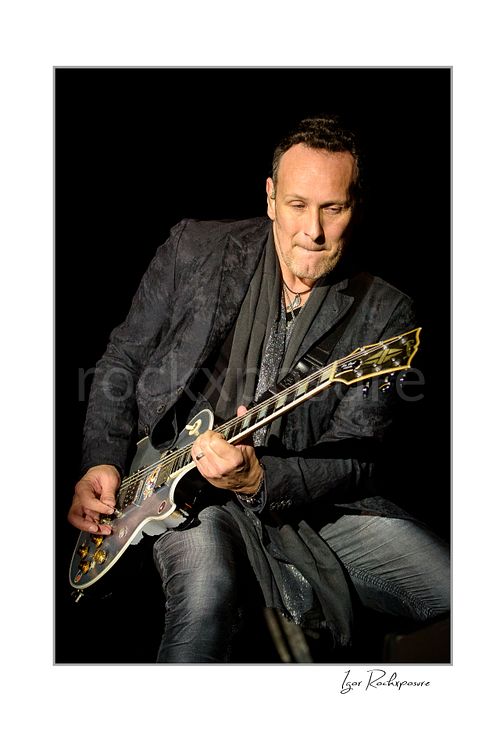 Vertical color image of Vivian Campbell of Def Leppard performing live on stage, playing electric guitar against a dark background