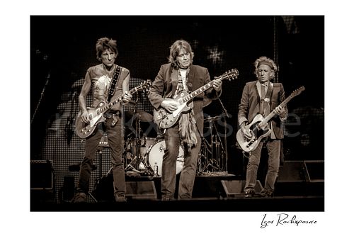 Horizontal black and white image of Ronnie Wood, Mick Taylor and Keith Richards of The Rolling Stones performing on stage with guitars