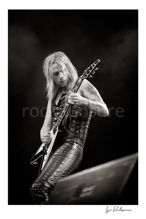 Vertical black and white image of Richie Faulkner of Judas Priest performing live on stage, playing flying V guitar against a dark background