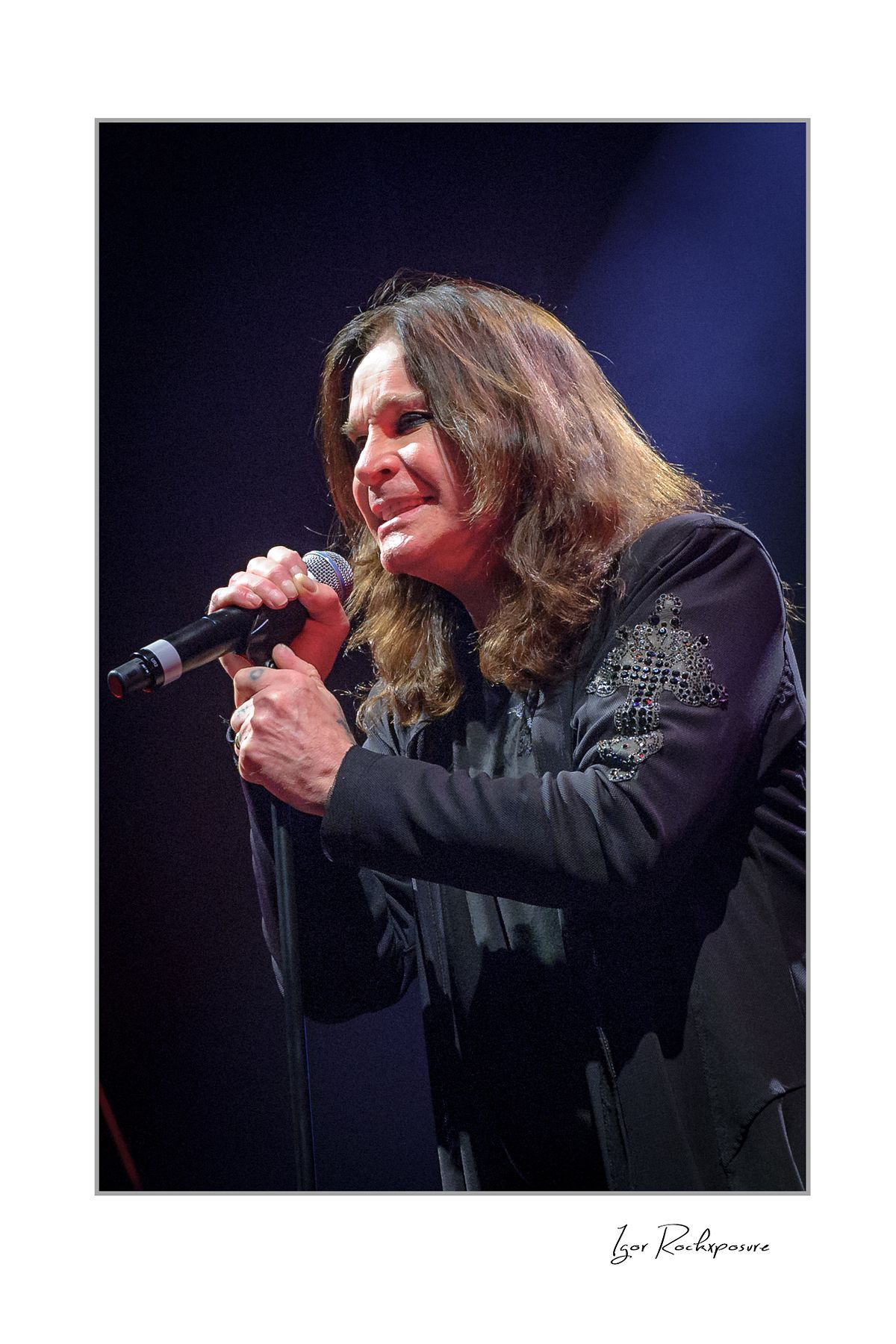 Ozzy Osbourne of Black Sabbath singing into a microphone during a live performance under soft blue concert lighting