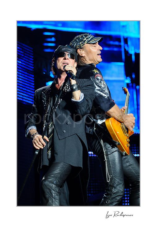 Vertical color image of Klaus Meine and Rudolf Schenker of Scorpions performing live together with microphone and orange guitar under blue stage lights