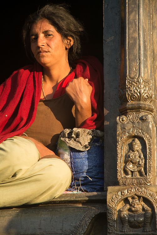 A woman with a weathered expression sits at sunset, bathed in light against a Nepalese column in a holy place for cremations in Kathmandu.