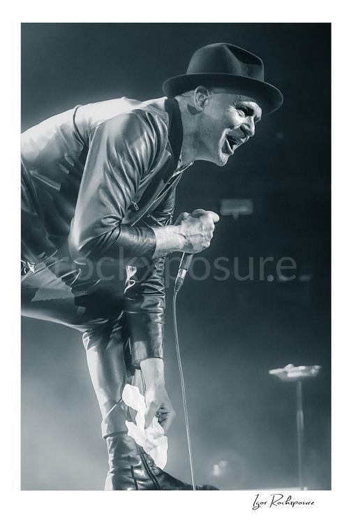 Vertical black and white image of Gord Downie of The Tragically Hip leaning forward and singing into a microphone, wearing a hat and leather jacket with a white cloth in hand amid bright concert haze