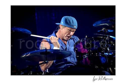 Horizontal color image of Chad Smith of Red Hot Chili Peppers playing drums in a focused live performance close-up, with drumstick raised above a purple drum kit under deep blue stage light