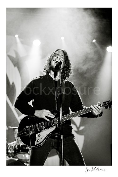 Vertical black and white image of Chris Cornell performing live on stage, singing into a microphone while playing electric guitar under bright concert lights