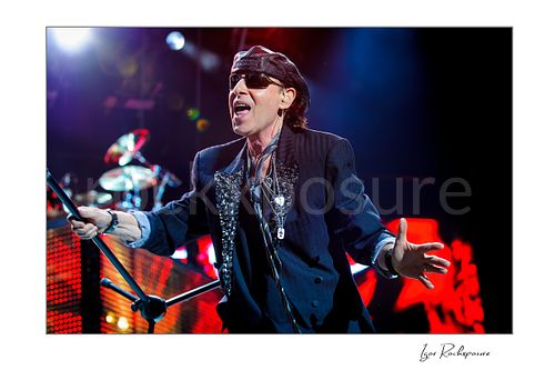 Horizontal color image of Klaus Meine of Scorpions performing live in sunglasses and cap with microphone stand under bright stage lights