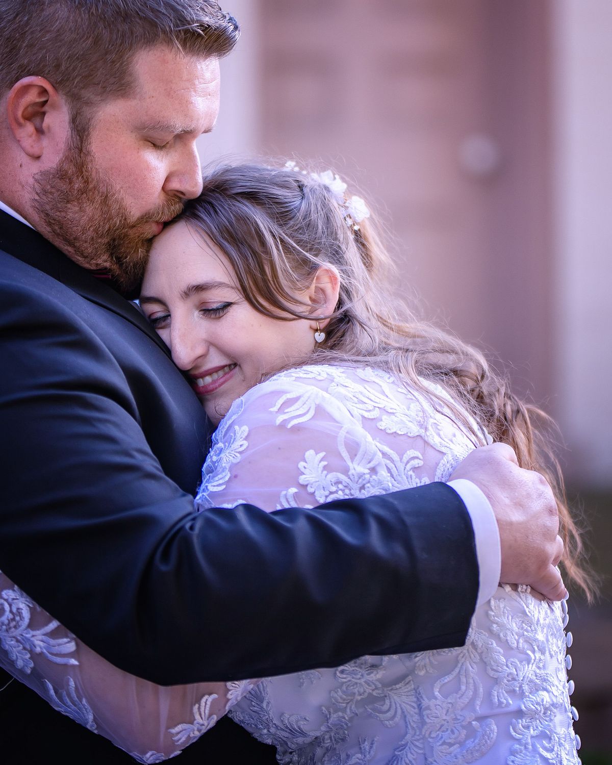 warm embrace between groom and bride, bride is closing her eyes and smiling, the groom is kissing her on her forehead.
