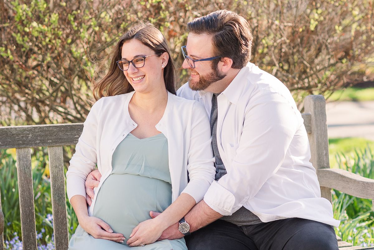 Husband and pregnant wife sit on bench in The Frick garden