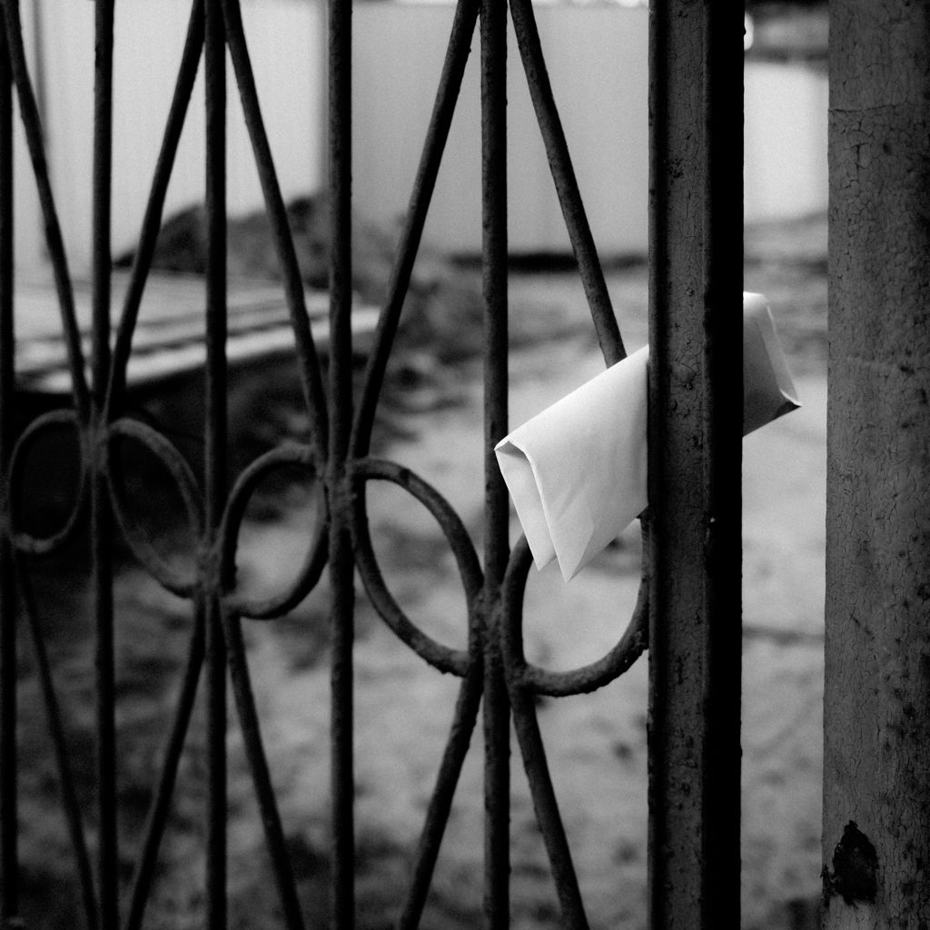 Czarno-biała fotografia autorstwa Michała Sumki przedstawiająca kopertę zawieszoną na metalowej bramie w Warszawie.