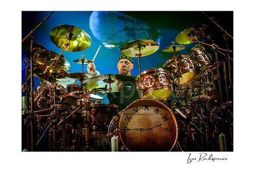Horizontal color image of Neil Peart performing behind an expansive drum kit during a Rush concert, surrounded by cymbals and toms with a raised drumstick under blue stage light