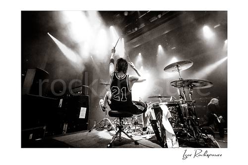 Horizontal black and white image of Zoltan Chaney playing drums from behind on stage under bright concert lights
