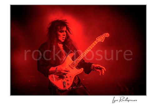 Horizontal color image of Yngwie Malmsteen performing live with a white guitar amid dense red stage smoke, illuminated by vivid red concert light