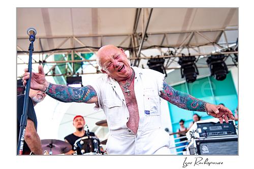 Horizontal color image of Angry Anderson of Rose Tattoo performing onstage in a white sleeveless outfit with arms outstretched and the drummer visible behind him