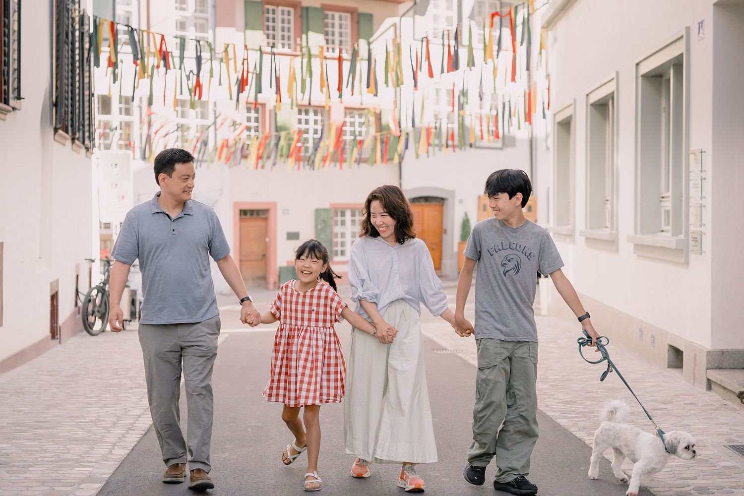 Basel natural light outdoor photographer capturing a joyful family walking hand in hand down a quaint, cobblestone street, with colorful streamers above. A small white dog on a leash joins them. The family is happy and enjoying every moment together.