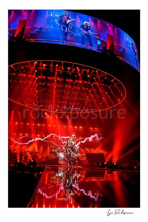 Vertical color image of Queen performing onstage beneath a massive circular lighting rig and giant overhead screen under vivid red concert light