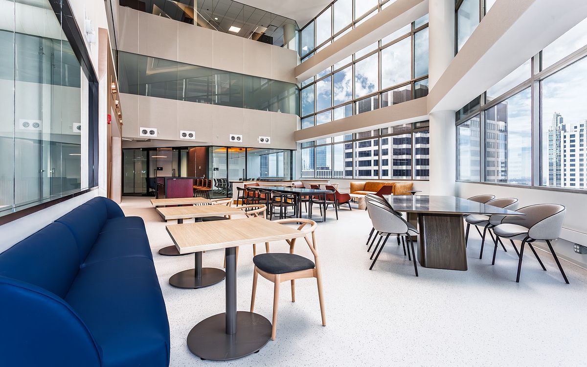 A bright, expansive three-story office atrium at 1 South Wacker in Chicago with a large blue velvet curved sofa, light wood cafe tables, and floor-to-ceiling windows overlooking the city.