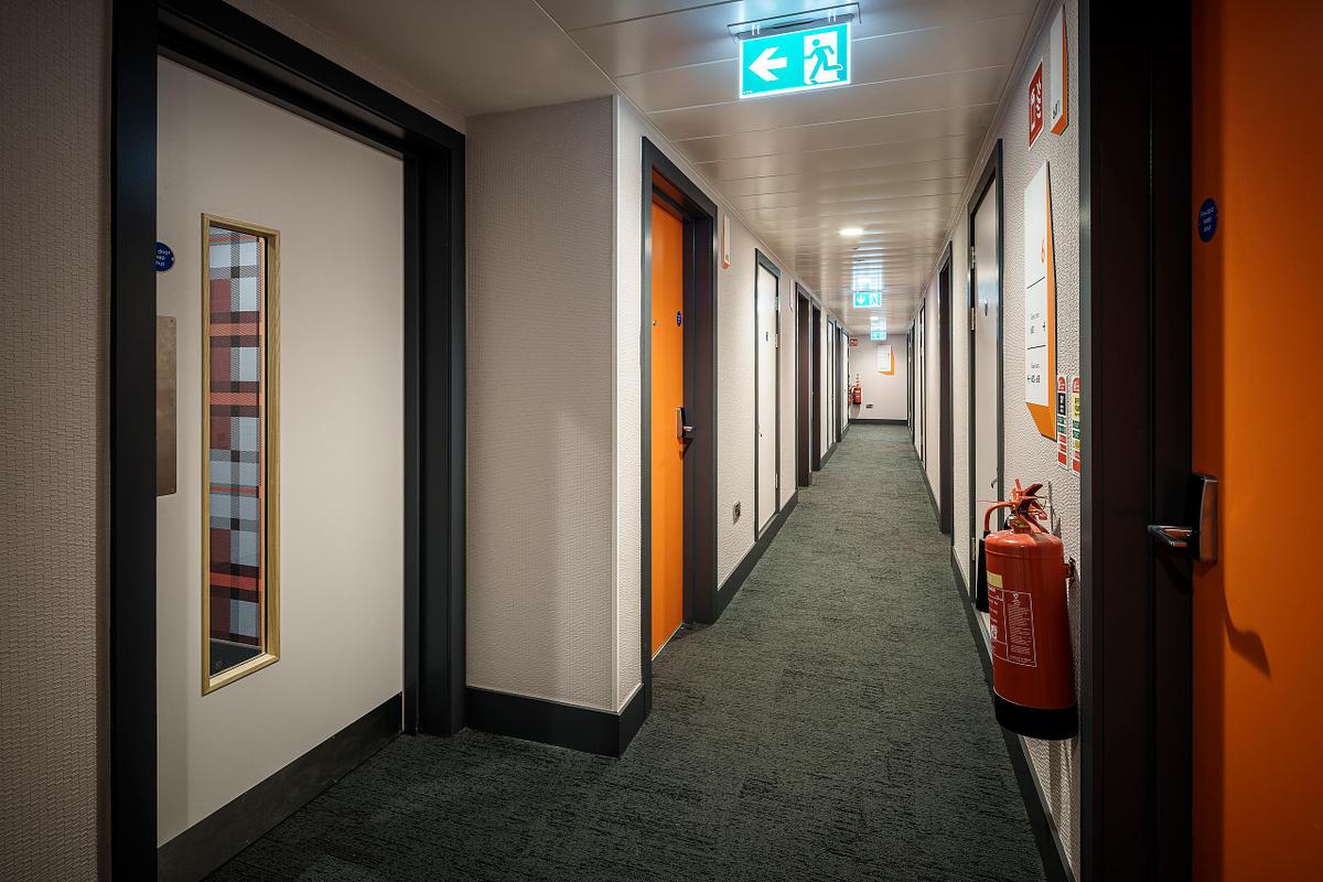Hotel corridor at the Easy Hotel Dublin. Multiple room doors, some with orange frames, are visible along the carpeted hallway. A fire extinguisher is mounted on the wall. An illuminated exit sign is on the ceiling.