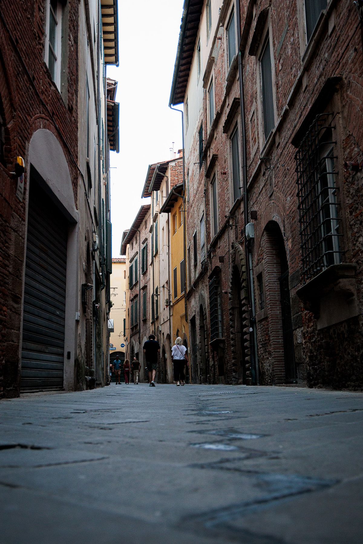 Low-angle view of a narrow cobblestone street in Lucca, Italy, lined with tall old brick and stone buildings in warm tones, including a yellow facade, with a few people walking in the distance and soft evening light creating a moody, historic atmosphere.