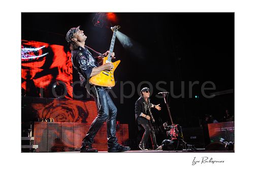 Horizontal color image of Rudolf Schenker and Klaus Meine of Scorpions performing live with orange guitar, microphone stand, and glowing red stage screens