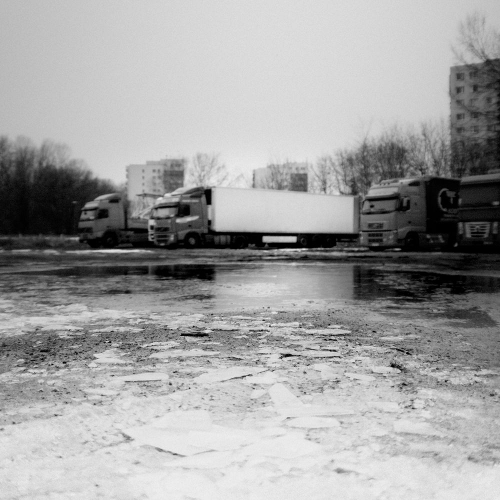 Czarno-biała fotografia autorstwa Michała Sumki ukazująca pierwszy mróz w Warszawie. Na pierwszym planie zamarznięta ziemia, a w tle zaparkowane ciężarówki i zabudowa miejska. Surowa, minimalistyczna kompozycja oddająca chłodny klimat miejskiego krajobrazu.