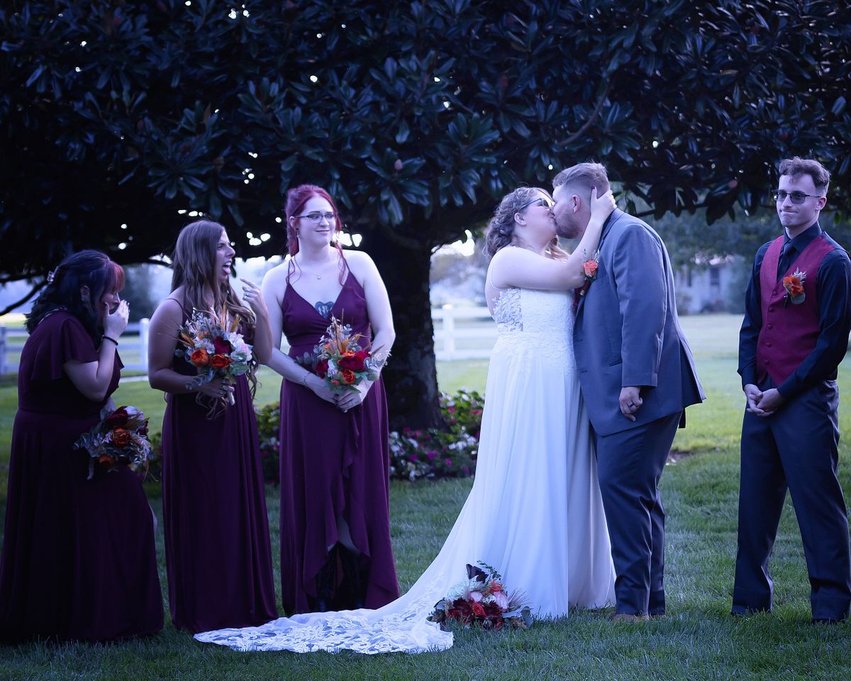 A wedding party at Willowbrook during blue hour, featuring a bride and groom kissing in front of a large tree in soft, natural evening light.