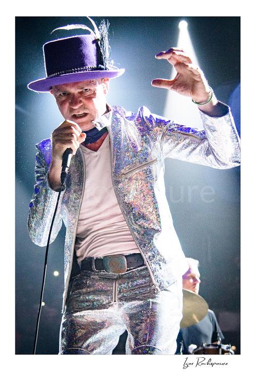 Vertical color image of Gord Downie of The Tragically Hip singing into a microphone in a dramatic close live performance shot, wearing a glittering silver suit and hat with bright stage light behind him