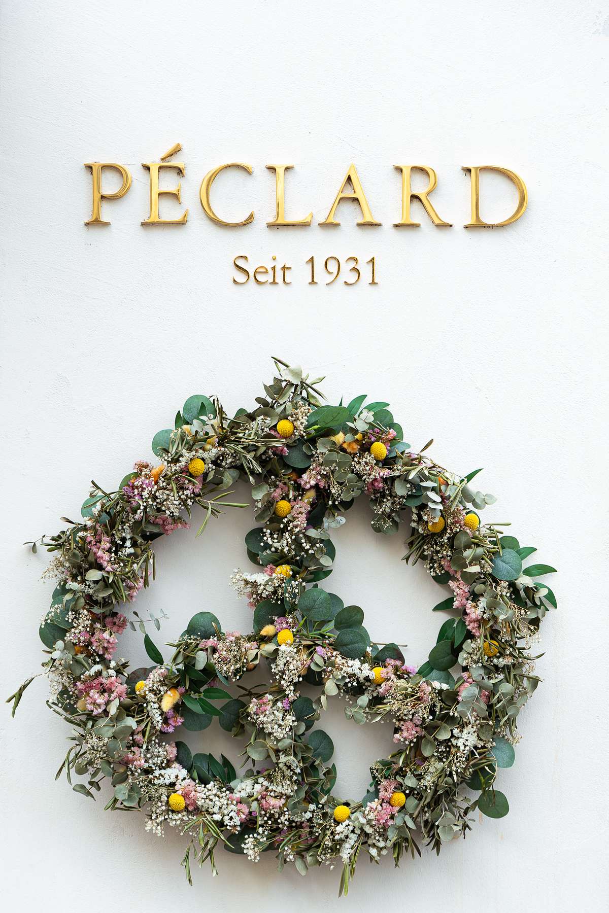 A circular wildflower wreath with eucalyptus leaves, white baby's breath, pink flowers, and yellow billy buttons hanging on a white wall beneath golden 'PECLARD Seit 1931' lettering at a historic Swiss shop in Zurich.