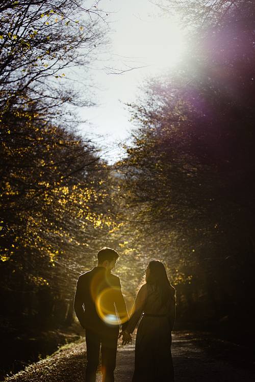 Coppia che cammina mano nella mano su un sentiero nel bosco marchigiano al tramonto, luce dorata e atmosfera romantica.