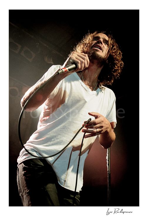 Vertical color image of Chris Cornell performing live on stage, holding a microphone under warm concert lighting