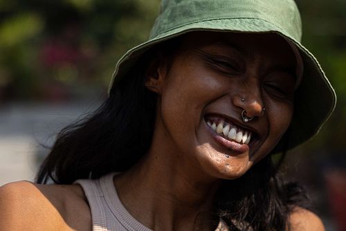 A headshot of an Indian woman smiling, with a nose ring, wearing a green bucket hat on a sunny day.
