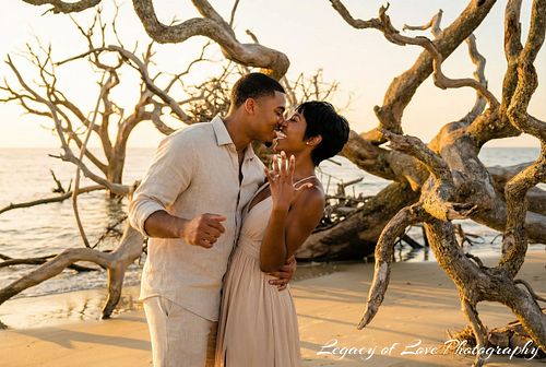 Couple embracing by driftwood trees during a golden hour sunset engagement session by Legacy of Love Photography.