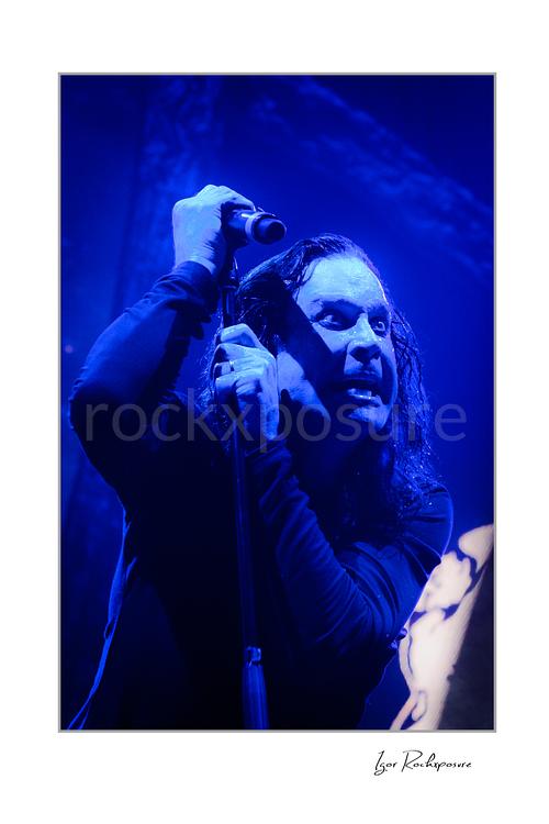 Ozzy Osbourne of Black Sabbath holding a microphone above his head during a live performance under deep blue concert lighting