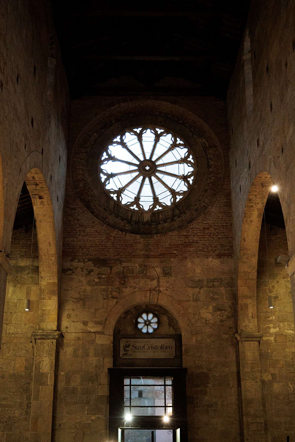 Interior view of Museo San Cristoforo in Lucca, Italy, showing a high stone wall with a large gothic circular window at the top, a smaller round floral window below and a doorway framed by a sign reading &ldquo;Museo San Cristoforo&rdquo;, with soft natural and artificial light revealing the rough, aged stone texture and creating a quiet, atmospheric mood.