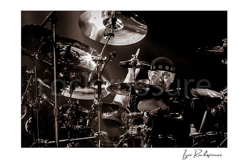 Horizontal black and white image of Neil Peart performing at his drum kit during a Rush concert, framed by cymbals and dramatic stage contrast