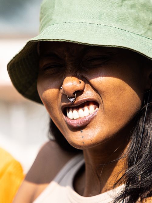 A headshot of an Indian woman smiling, with a nose ring, wearing a green bucket hat on a sunny day.