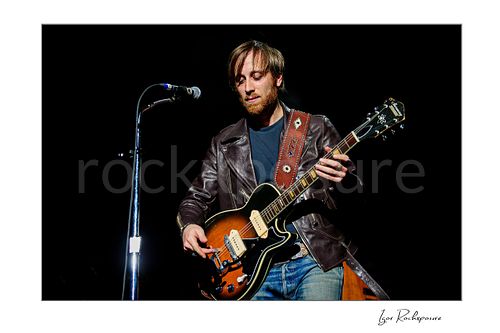 Horizontal color image of Dan Auerbach of The Black Keys playing guitar beside a microphone in a dark concert scene