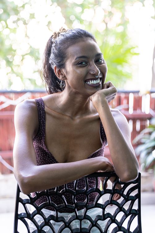 A portrait of an Indian woman, smiling and wearing a purple top, sitting on a chair on a balcony with her arms placed on the backrest.