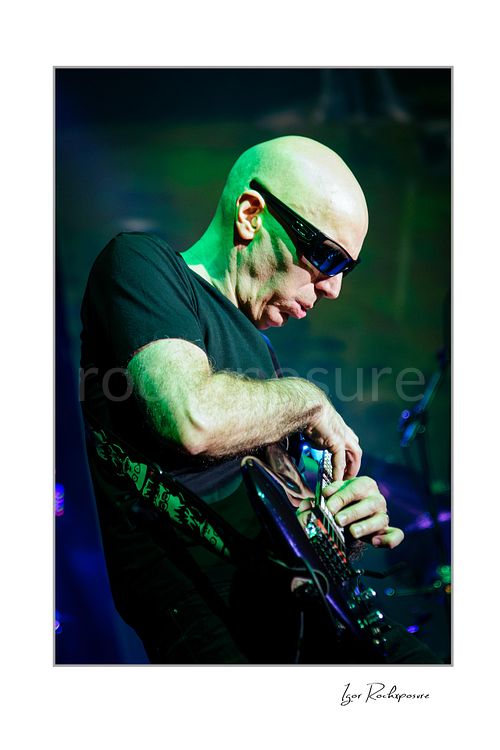 Vertical color image of Joe Satriani performing live in sunglasses with black guitar and vivid green stage lighting