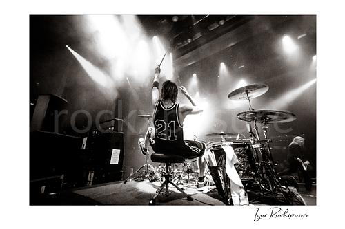 Horizontal black and white image of Zoltan Chaney playing drums from behind on stage under bright concert lights