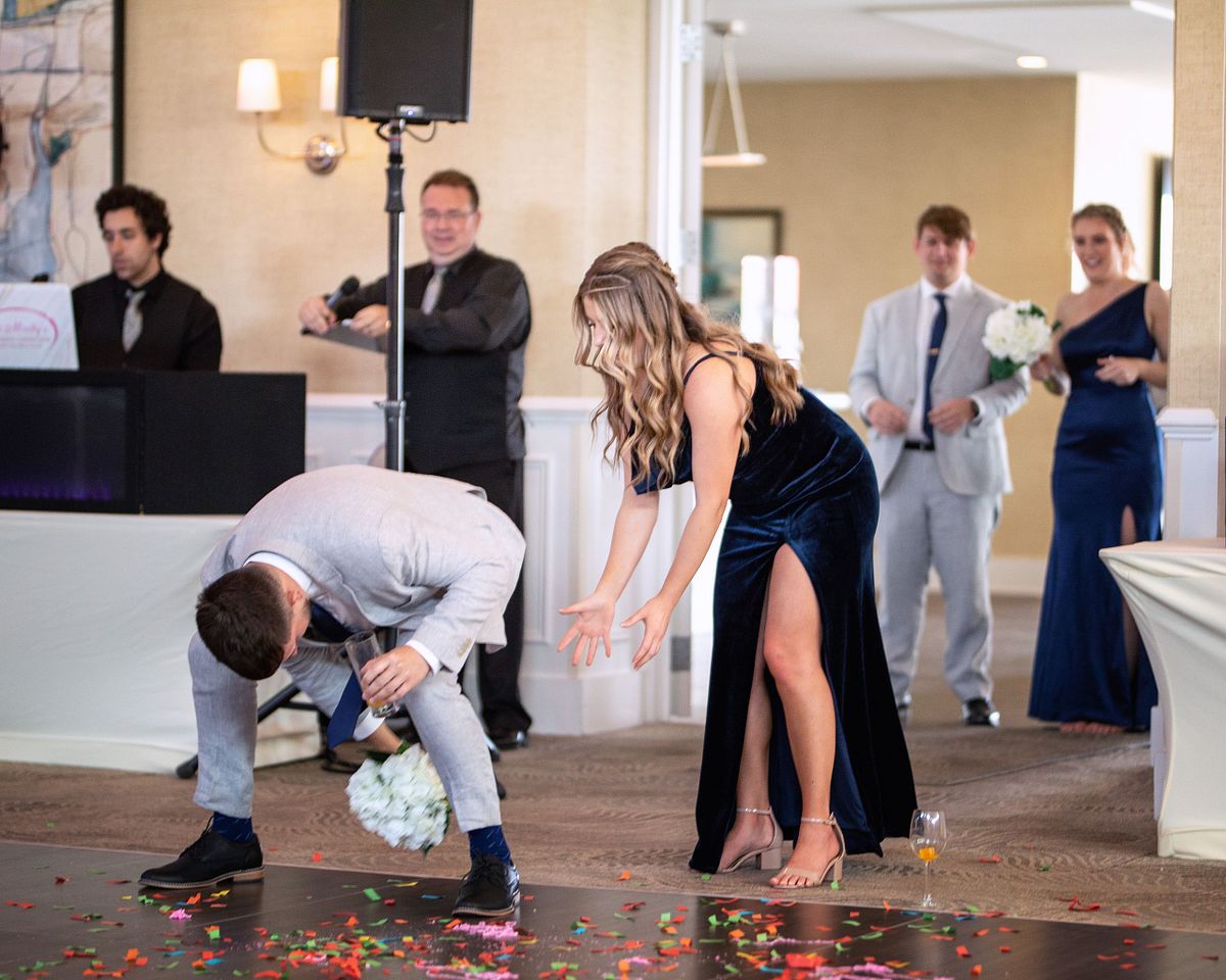 bridesmaid and groomsmen entering the ball room, the groomsmen is tossing a flower bouquet to the bridesmaid, he is wearing a grey suite, she is wearing a blues dress.