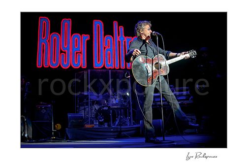 Horizontal color image of Roger Daltrey performing with a guitar onstage beside a microphone with his name glowing on the backdrop behind him