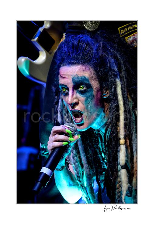 Vertical color image of Calico Cooper of Beasto Blanco singing into a microphone in an intense live performance close-up, with dramatic eye makeup, dreadlocks, and neon green nails under blue stage light
