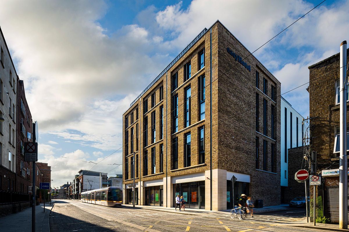 Street-level view of the Easy Hotel Dublin, showing its modern brick facade and surrounding urban environment. A tram is passing on the street, and pedestrians are visible. The 'easyHotel Dublin City Centre' sign is on the building. A Lycamobile shop is on the right commercial side of the building.